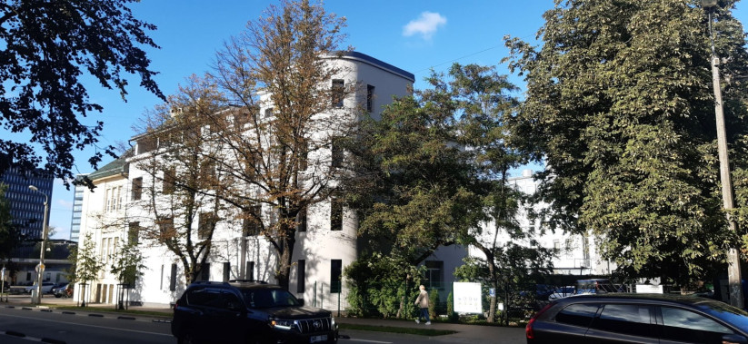 Ēkas fasāde ar kokiem priekšplānā, saulainā dienā.