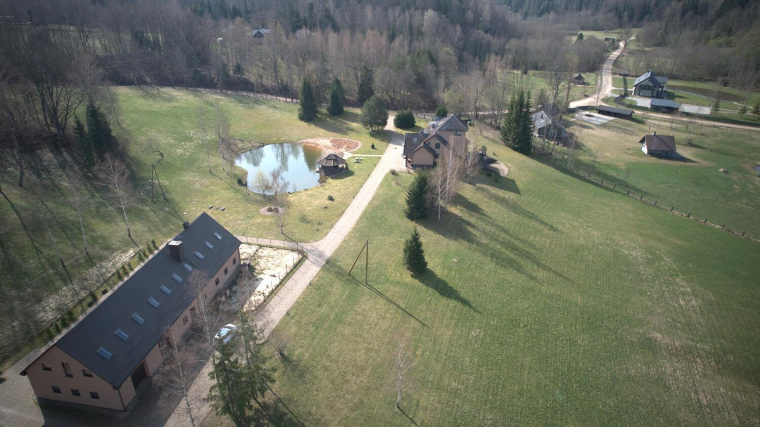 Aeriāla fotogrāfija ar māju un dīķi zaļā ainavā.