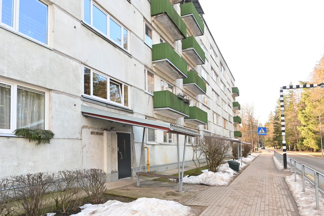 Piecu stāvu māja ar zaļiem balkoniem atrodas pie asfaltētas ielas un gar to iet bruģēta ietve