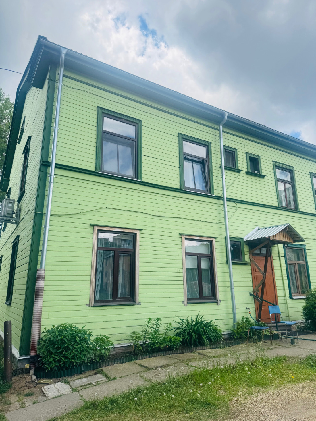 Zaļas koka mājas fasāde ar brūnām durvīm un soliņu.