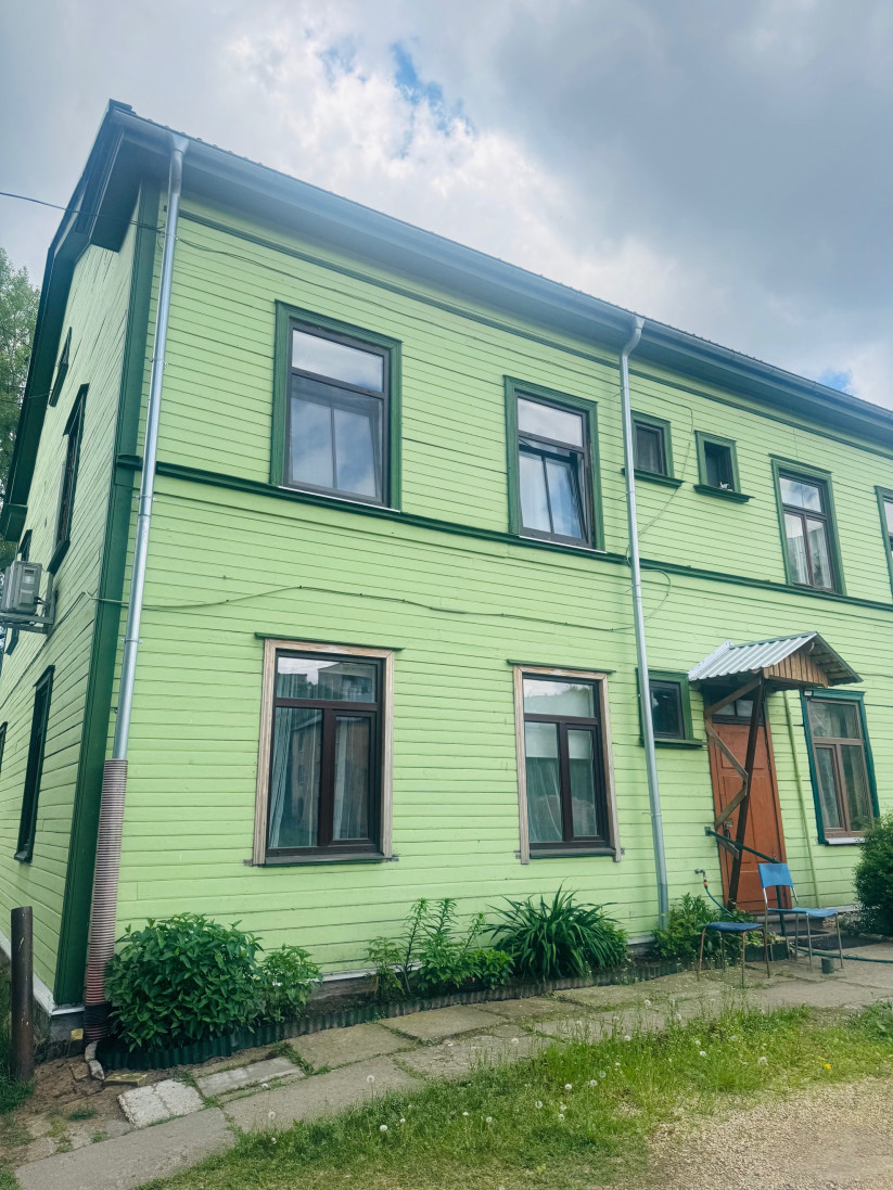 Zaļas koka mājas fasāde ar brūnām durvīm un soliņu.